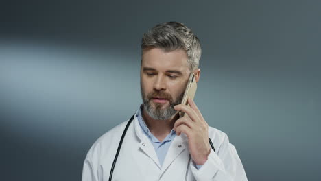 Männlicher-Arzt,-Der-Auf-Dem-Handy-Spricht-Und-Beratung-Durch-Verbindung-Auf-Dem-Grauen-Wandhintergrund-Durchführt
