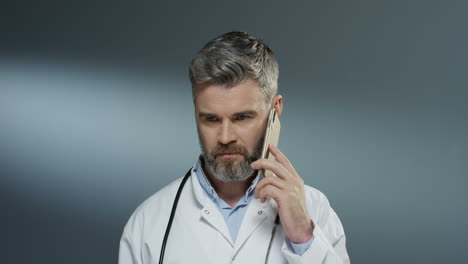 Gut-Aussehender-Grauhaariger-Männlicher-Sanitäter-Im-Weißen-Kleid,-Der-Auf-Dem-Handy-Auf-Dem-Grauen-Hintergrund-Spricht
