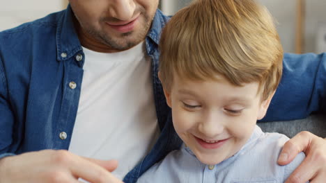Close-Up-Of-The-Cute-Small-Boy-Sitting-Next-To-His-Good-Looking-Father,-Playing-A-Game-On-The-Smartphone-And-Smiling