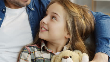 Close-Up-Of-The-Teenage-Cute-Blonde-Girl-Sitting-And-Hugging-Her-Teddy-Bear-While-Her-Father-Hugging-Her
