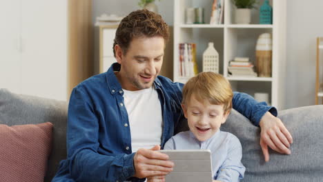 Handsome-Man-Sitting-On-The-Gray-Sofa-And-Hugging-His-Cute-Little-Son-When-They-Watching-Something-On-The-Tablet-Computer-And-Talking-In-The-Living-Room