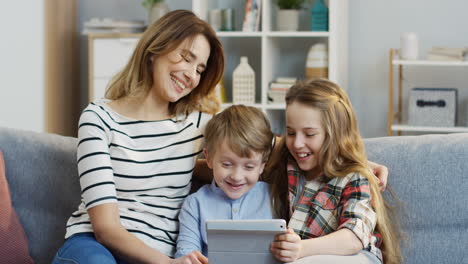 Portrait-Of-The-Pretty-Mother-Sitting-On-The-Gray-Couch-And-Hugging-Her-Cute-Small-Son-And-Teen-Daughter-While-They-Watching-Something-On-The-Tablet-Computer-In-The-Living-Room