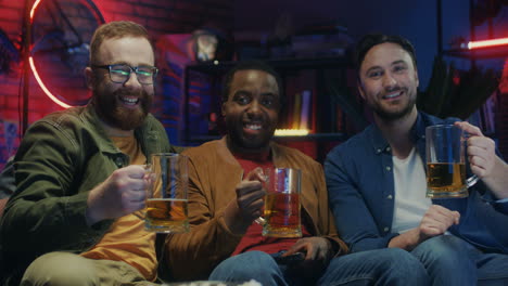 Three-Multiethnic-Young-Guys-Doing-Cheers-Gesture-With-Beer-And-Smiling-To-The-Camera-At-Home-At-Night-While-Watching-Tv