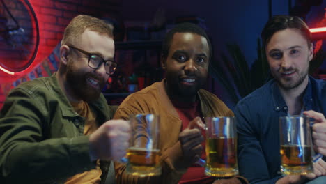 Mixed-Races-Young-Three-Men-Doing-Cheers-Gesture-With-Beer-And-Smiling-Cheerfully-To-The-Camera-In-The-Dark-Room-While-Watching-Sport-Game-On-Tv