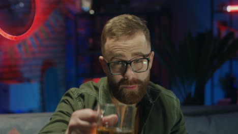 Portrait-Of-The-Young-Cheerful-Man-In-Glasses-Drinking-Beer-And-Doing-Cheers-Gesture-To-The-Camera-And-Smiling-In-The-Dark-Room