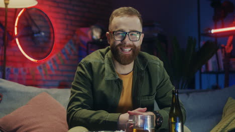 Portrait-Of-The-Joyful-Red-Haired-Young-Man-In-Glasses-Smiling-To-The-Camera-And-Being-In-Good-Mood-While-Sitting-On-The-Sofa-In-The-Dark-Room