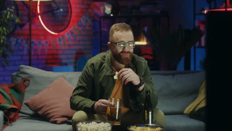 Young-Guy-In-Glasses-Sitting-On-The-Couch-With-Beer-And-Snacks-An-Worrying-About-Sport-Match-On-Tv-At-Night