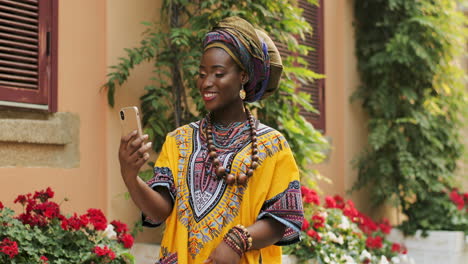 Portrait-Shot-Of-The-Young-Beautiful-Woman-In-The-Traditional-Clothes-Standing-In-The-Nice-Courtyard-With-Flowers-And-Having-Videochat-On-The-Smartphone