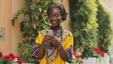Young-Charming-Smiled-Woman-In-The-Traditional-Clothes-Standing-In-The-Nice-Courtyard-With-Flowers,-Typing-And-Texting-On-The-Smartphone