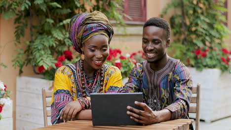 Cheerful-Young-Stylish-Man-And-Woman-In-Traditional-Outfits-Laughing-While-Sitting-At-The-Table-In-The-Nice-Yard-With-Flowers-And-Watching-Something-On-The-Tablet-Computer