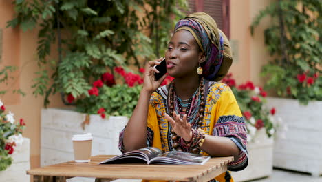 Pretty-Young-Woman-In-The-Traditional-Outfit-Talking-On-The-Phone-Cheerfully-While-Sitting-At-The-Table-Wit-Magazine-And-Coffee-In-The-Beautiful-Courtyard