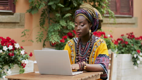 Good-Looking-Young-Woman-In-The-Traditional-Clothes-Working-On-The-Laptop-Computer,-Tapping-And-Texting,-While-Sitting-At-The-Table-In-The-Nice-Yard-Outdoor