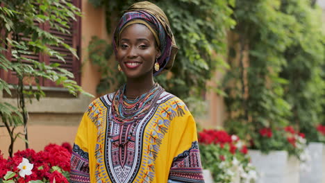 Porträtaufnahme-Der-Schönen-Und-Glücklichen-Afrikanischen-Frau-Im-Traditionellen-Outfit,-Die-Im-Gemütlichen-Innenhof-Mit-Blumen-Steht-Und-In-Die-Kamera-Lächelt