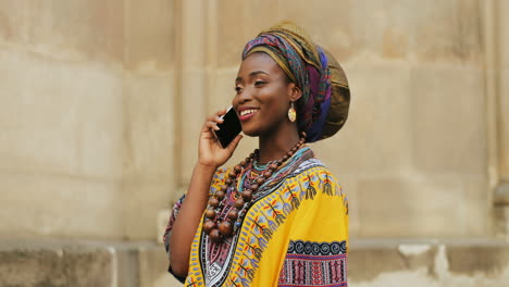 Beautiful-Young-Charming-Woman-In-The-Traditional-Outfit-And-With-Scarf-On-The-Head-Talking-Joyfully-On-The-Phone-On-The-Wall-Background