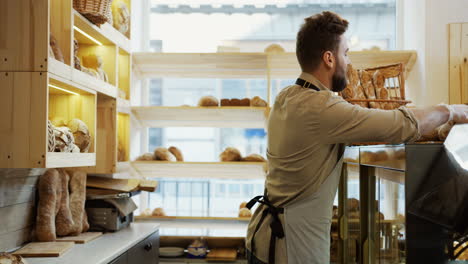 Good-Looking-Young-Male-Baker-Carrying-New-Portion-Of-The-Fresh-Bread-To-The-Counter-In-The-Morning