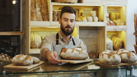 Gutaussehender-Junger-Bäckereiverkäufer,-Der-Morgens-Vor-Der-Eröffnung-Frisches-Brot-Auf-Die-Theke-Riecht-Und-Legt
