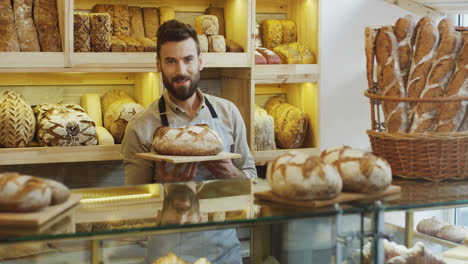 Gut-Aussehender-Junger-Männlicher-Bäckereiverkäufer,-Der-Morgens-Frisches-Brot-An-Der-Theke-Riecht-Und-Vor-Der-Kamera-Posiert