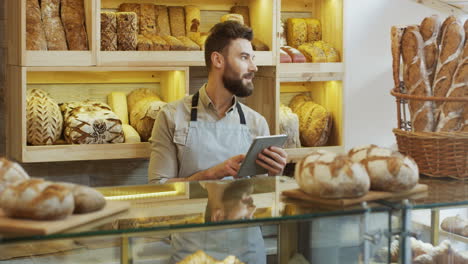 Handsome-Male-Bread-Seller-Typing-On-The-Tablet-Device-At-The-Counter-In-The-Bakery-Shop,-Then-Smiling-To-The-Camera