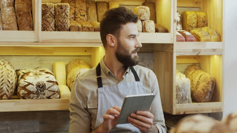 Attractive-Man-Bread-Seller-Using-His-Tablet-Computer-While-Standing-At-The-Counter-In-The-Bakery-Shop,-Then-Looking-At-The-Side-In-The-Window