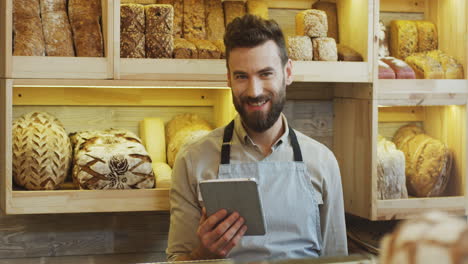 Young-Man-Bread-Seller-Scrolling-And-Taping-On-The-Tablet-Computer-While-Standing-At-The-Counter-In-The-Bakery-Shop,-Then-Looking-To-The-Camera