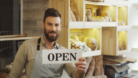Male-Young-Bakery-Seller-Posing-With-A-Signboard-Open-Inside-Of-The-Shop-Of-Bread