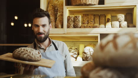 Portrait-Shot-Of-The-Young-Male-Baker-Looking-At-The-Side-While-Holding-A-Tray-With-Bread-Then-Looking-At-The-Camera-In-The-Shop