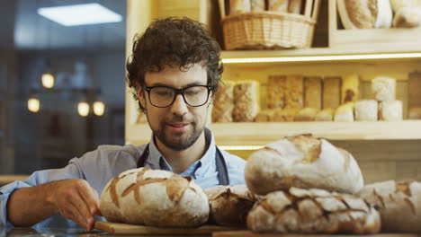 Gut-Aussehender-Mann,-Bäckereiverkäufer-In-Gläsern,-Der-Morgens-Frisches-Brot-Auf-Die-Theke-Bringt-Und-Daran-Riecht