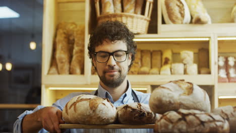 Porträtaufnahme-Des-Gutaussehenden-Männlichen-Bäckereiverkäufers-Mit-Brille,-Der-Frisches-Brot-Auf-Die-Theke-Legt-Und-In-Die-Kamera-Lächelt