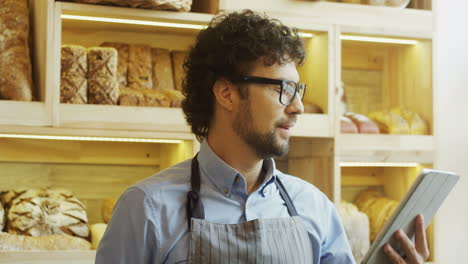 Handsome-Man-Bread-Seller-In-Glasses-Using-His-Tablet-Computer-While-Standing-At-The-Counter-In-The-Bakery-Shop