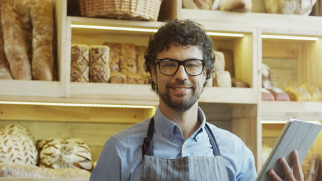 Attractive-Male-Bread-Vendor-In-Glasses-Scrolling-And-Taping-On-The-Tablet-Device-While-Standing-At-The-Counter-In-The-Bakery-Shop,-Then-Smiling-To-The-Camera