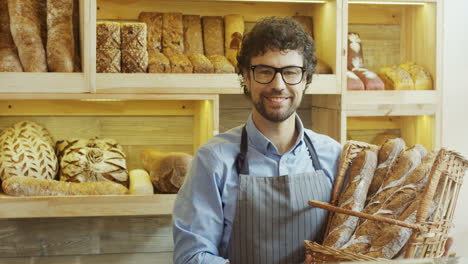 Porträtaufnahme-Des-Gutaussehenden-Männlichen-Bäckers,-Der-Lächelt,-Während-Er-Mit-Baguettes-Im-Korb-Im-Laden-Posiert