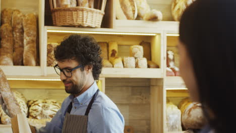 Close-Up-Of-The-Attractive-Male-Bakery-Seller-Giving-Baguettes-To-The-Young-Woman-When-She-Buying-Them
