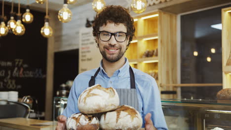 Portrait-Of-The-Young-Man-Baker-Holding-A-Tray-With-Bread-Then-Looking-At-The-Camera-In-The-Shop