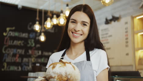 Close-Up-Of-The-Beautiful-Young-Female-Baker-Looking-At-The-Camera-While-Holding-Fresh-Bread-In-The-Shop