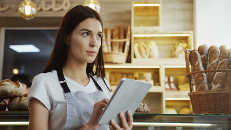Pretty-Female-Baker-In-Glasses-Scrolling-And-Taping-On-The-Tablet-Computer-While-Standing-At-The-Counter-In-The-Shop