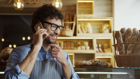 Male-Seller-In-Glasses-Talking-Cheerfully-On-The-Phone-While-Standing-At-The-Counter-In-The-Bakery-Shop