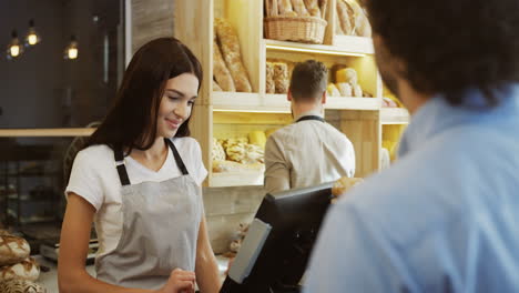 Young-Beautiful-Woman-Standing-At-The-Counter-In-The-Bakery-Shop-And-Selling-Bread-To-The-Man,-Her-Male-Co-Worker-Doing-Something-Behind-Her