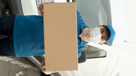 Portrait-Of-Young-Handsome-Deliveryman-In-Medical-Mask-And-Mail-Uniform-With-Cap-Standing-Outdoor-At-Car-And-Handing-Carton-Box-To-Camera