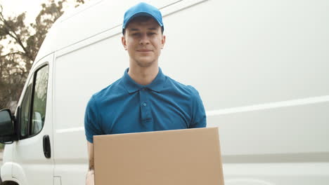 Portrait-Of-Young-Handsome-Deliveryman-In-Blue-Uniform-With-Cap-Standing-Outdoor-At-Car-And-Handing-Carton-Box-To-Camera