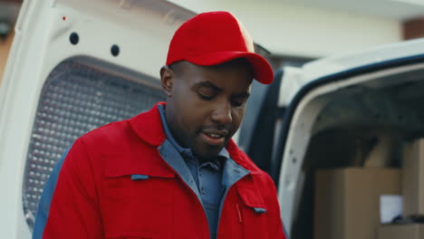 Close-Up-Of-The-Young-Deliveryman-In-The-Red-Uniform-And-Cap-Standing-Outdoor-At-The-White-Van-And-Counting-Mail-Boxes
