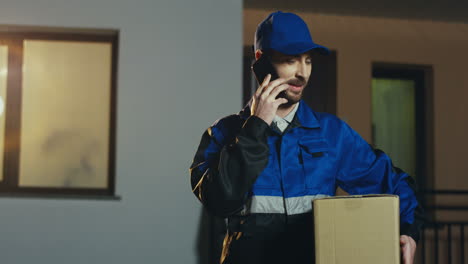 Attraktiver-Postbote-Im-Blauen-Kostüm-Und-Kappe-Mit-Kartonpercel,-Der-Am-Handy-Spricht