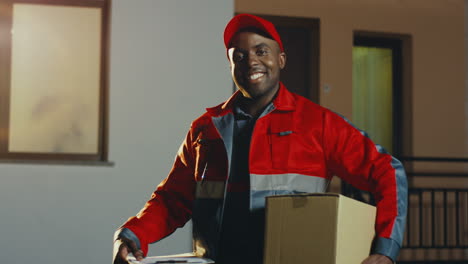 Portrait-Of-The-Young-And-Cheerful-Mailman-In-The-Red-Costume-And-A-Cap-With-A-Box-Looking-At-The-Side-And-Then-Turning-His-Head-To-The-Camera-In-The-Evening