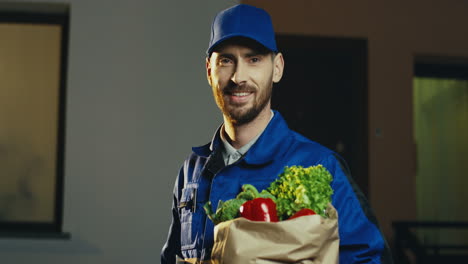 Primer-Plano-Del-Atractivo-Trabajador-Masculino-De-La-Entrega-Del-Supermercado-Posando-Y-Sonriendo-A-La-Cámara-Con-Un-Paquete-De-Cartón-Lleno-De-Verduras-Frescas