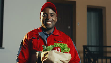 Portrait-Of-The-Young-Smiled-Male-Worker-Of-The-Supermarket-Delivery-Posing-To-The-Camera-With-A-Carton-Package-Full-Of-Fresh-Vegetables-In-The-Evening