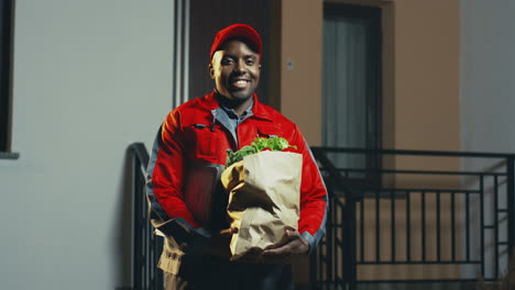 Portrait-Shot-Of-The-Young-Man-From-The-Delivery-Service-Of-Supermarket-In-The-Red-Costume-And-Cap-With-A-Fresh-Vegetables-In-The-Carton-Package-Looking-At-The-Side-And-Then-Smiling-To-The-Camera-At-Night