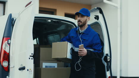 Young-Handsome-And-Joyful-Mailman-In-Blue-Uniform-Taking-Out-Carton-Box-From-A-Van-And-Walking-To-The-House-While-Listening-To-The-Music-In-Headphones-And-Smiling