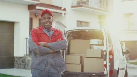Joyful-Young-Deliveryman-In-The-Red-Uniform-And-Cap-Standing-At-The-White-Male-Van-With-Boxes,-Crossing-Hands-In-Front-Of-Him-And-Smiling-To-The-Camera