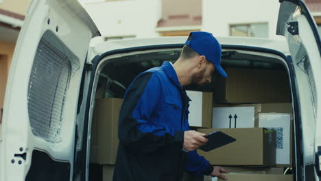 Good-Looking-Mailman-In-Blue-Costume-And-Cap-Checking-The-Mail-Boxes-With-Tablet-Device-In-Hands