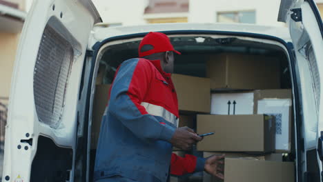 Young-Man-Worker-Of-Mail-Company-Counting-And-Checking-Parcels-In-The-Van-With-Tablet-Device-In-Hands