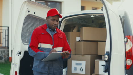 Postbote-In-Roter-Uniform-Und-Mütze-Steht-Mit-Kartons-Am-Weißen-Lieferwagen-Und-Füllt-Dokumente-In-Der-Zwischenablage-Aus
